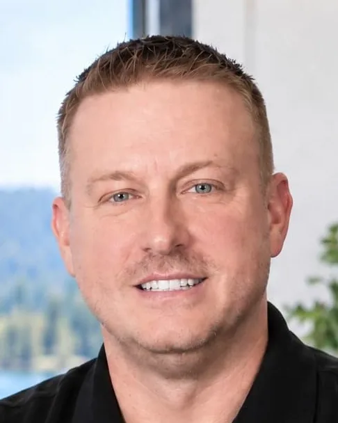 Jon Carey, founder of Carey Enterprises, smiling in a black polo at his desk in Athol, Idaho, with a North Idaho sign and a lake view behind him
